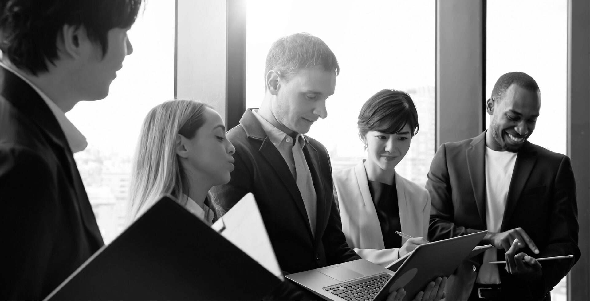 Multi-ethnic business team gathered near office window, reviewing information on laptop and documents